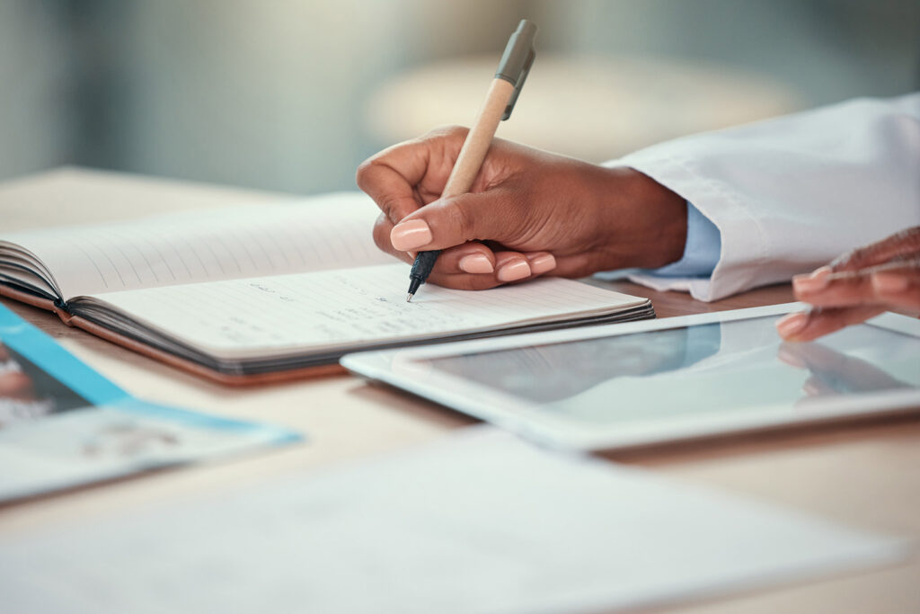 a close up image of a doctor writing notes while scrolling a tablet